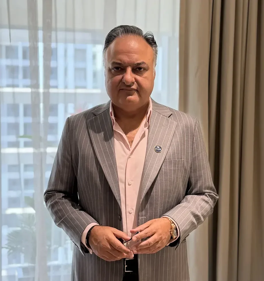 Professional portrait of a man in a gray pinstripe suit and pink shirt, standing by a window and holding glasses.