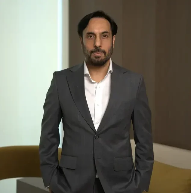 Man in a dark gray suit with a white shirt standing indoors, hands in pockets, looking at the camera in a neutral pose