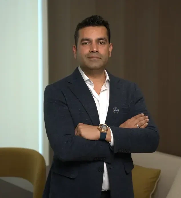 Man in a dark blazer with a white shirt, arms crossed, standing in a modern office. (informative, concise)